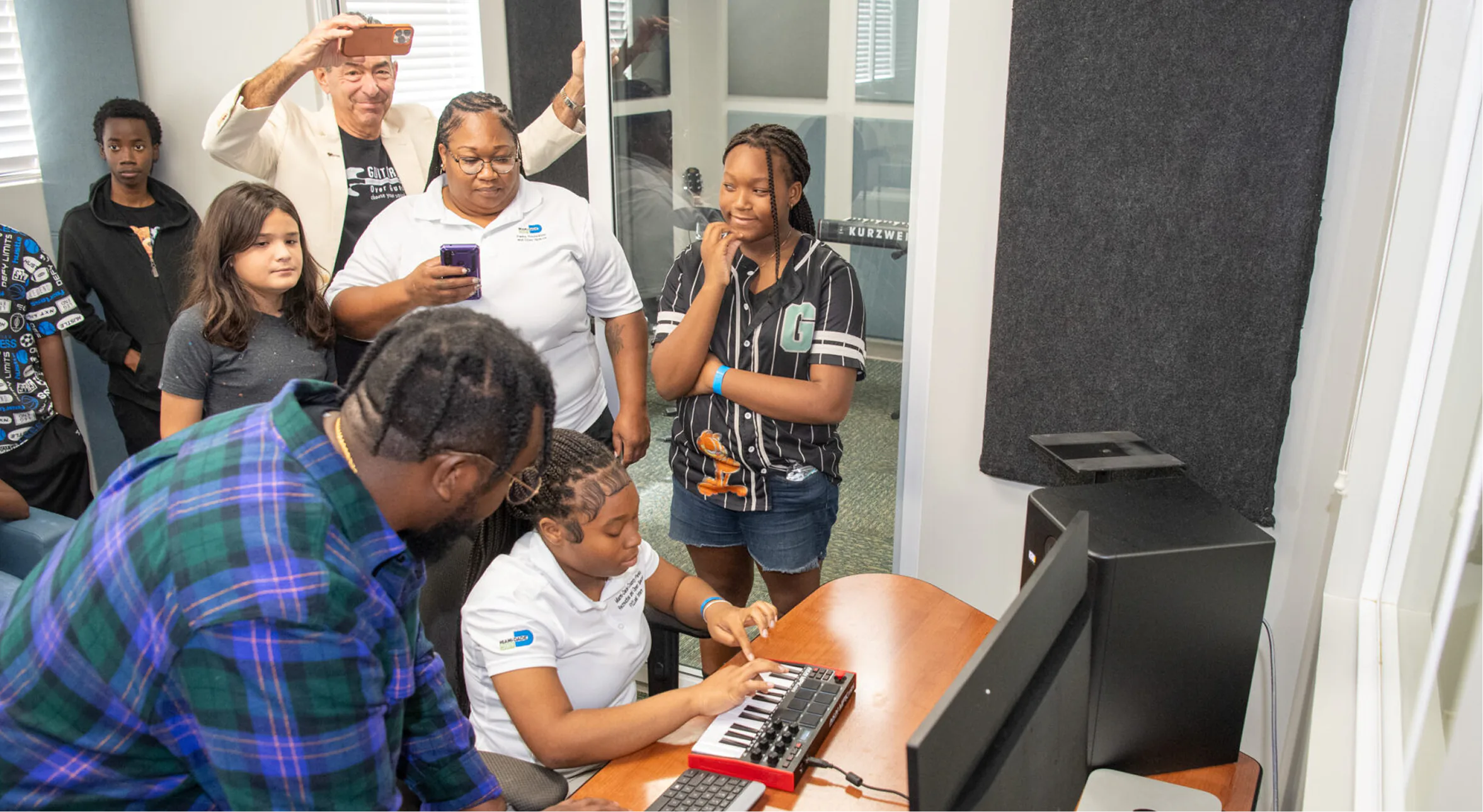 A music instructor helps a student play a MIDI keyboard while other students and an adult observe in a music studio.